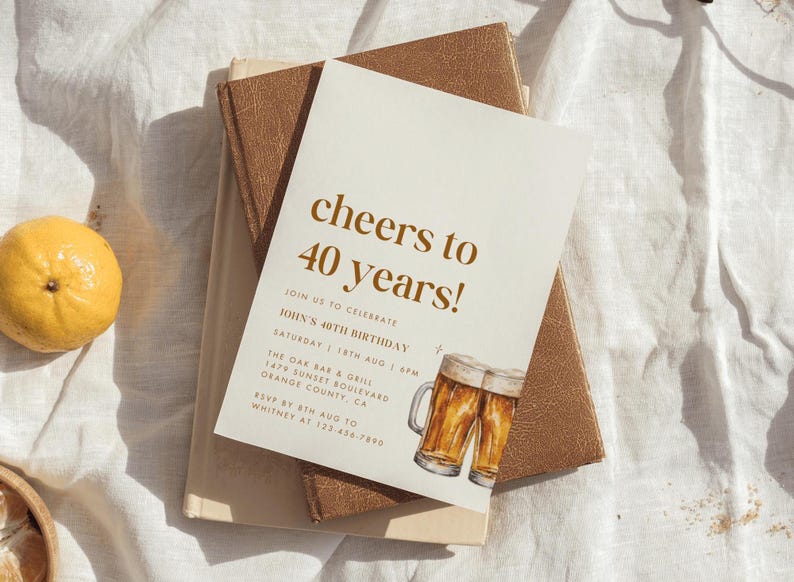 May include: A cream-colored invitation with the text "cheers to 40 years!" in brown lettering. The invitation includes details for John's 40th birthday, with watercolor illustrations of beer mugs. The invitation rests on a stack of books.