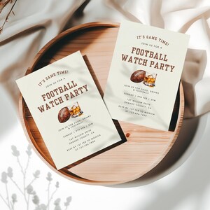 May include: A beige invitation card with brown text that reads "It's Game Time! Join us for a Football Watch Party". The card features a watercolor illustration of a football and a glass of whiskey. The text also includes the date, time, and location of the party.