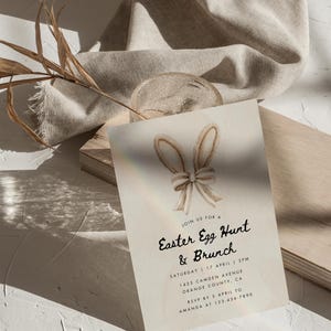 May include: A light beige invitation card with a watercolor illustration of bunny ears with a bow. The text on the card reads "Join us for a Easter Egg Hunt & Brunch, Saturday 17 April 2PM, 1425 Camden Avenue Orange County, CA, RSVP by 5 April to Amanda at 123-456-7890".