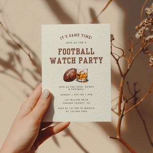 May include: A beige invitation card with brown text that reads "It's Game Time! Join us for a Football Watch Party". The card also includes a watercolor illustration of a football and a glass of whiskey.