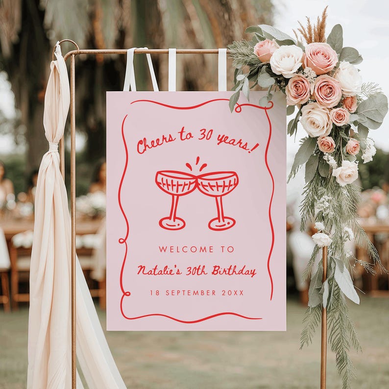 May include: A pink sign with red text and illustrations. The sign reads "Cheers to 30 years!" above two champagne glasses. Below, it says "Welcome to Natalie's 30th Birthday" and the date "18 September 20XX". The sign is part of a birthday celebration.