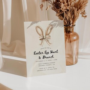May include: A light brown invitation card with a watercolor illustration of bunny ears with a bow. The text on the card reads "Join us for a Easter Egg Hunt & Brunch" with the date, time, and location details below.