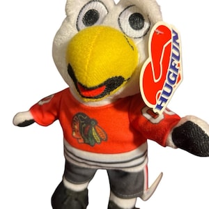 May include: A plush toy eagle with a white head, yellow beak, and black eyes. It wears a red jersey with a team logo and gray shorts. The tag reads "HUGFUN".
