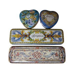 May include: An assortment of decorative metal boxes in various shapes. Two heart-shaped boxes with floral and ornate designs, and two rectangular boxes with the word "Brighton" and floral patterns. The boxes are in shades of blue, yellow, and white.