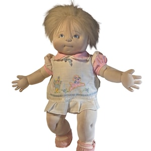 May include: A soft-sculpture doll with light brown yarn hair and blue eyes, wearing a white sleeveless dress with a pink collar, pink puff sleeves, and an embroidered design featuring a pink animal figure and a basket of colorful flowers.