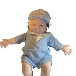 May include: A soft-sculpture doll with a round face, brown hair, and a light blue ribbed romper. The doll wears a matching light blue cap, a collared shirt underneath the romper, and white shoes with laces. The doll has an open-armed pose.