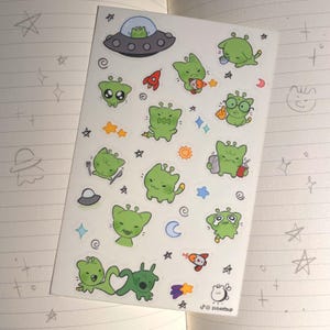 May include: A sticker sheet with green alien illustrations in various poses. The stickers include aliens in a spaceship, holding food, and with accessories. The background is a notebook page with doodles. The artist's name is visible.