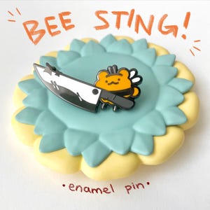 May include: Enamel pin featuring a cartoon bee holding a knife, with the text "BEE STING!" in orange. The pin is set on a light blue and yellow flower-shaped base. The words "enamel pin" are written in red.
