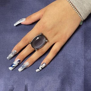 May include: A hand wearing a silver ring with a large, oval-shaped, dark gray gemstone. The hand also has long, white nails with blue and white floral designs.