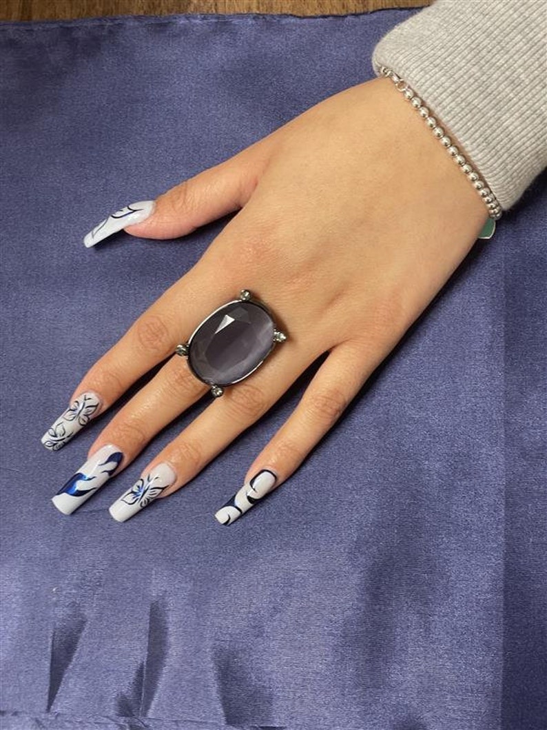 May include: A hand wearing a silver ring with a large, oval-shaped, dark gray gemstone. The hand also has long, white nails with blue and white floral designs.