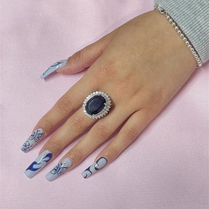 May include: A silver ring with a large oval-shaped blue gemstone surrounded by smaller clear gemstones. The ring is on a person's finger with blue and white nail polish.