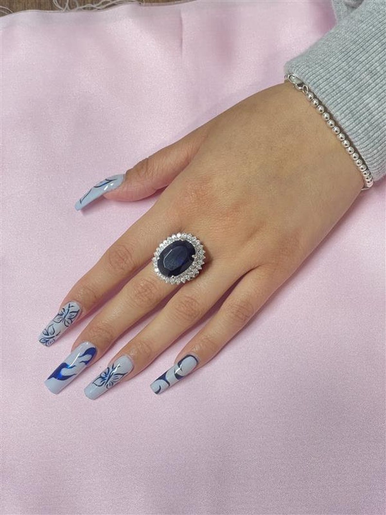 May include: A silver ring with a large oval-shaped blue gemstone surrounded by smaller clear gemstones. The ring is on a person's finger with blue and white nail polish.