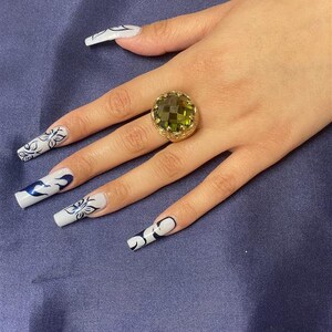 May include: A gold ring with a large, faceted, green gemstone. The ring is on a finger with white nail polish and black and white nail art.