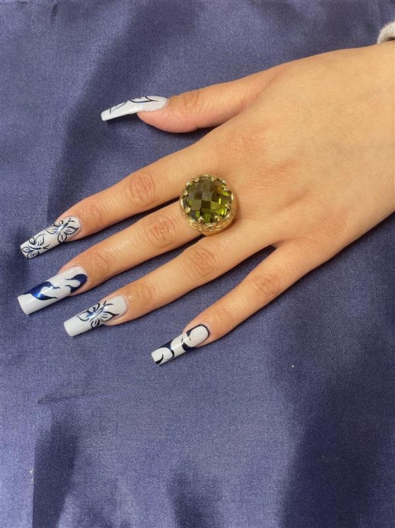 May include: A gold ring with a large, faceted, green gemstone. The ring is on a finger with white nail polish and black and white nail art.