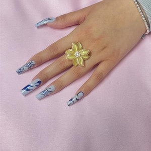 May include: A yellow flower-shaped ring with a clear rhinestone center. The ring is on a finger with light blue nail polish and a blue and white design.