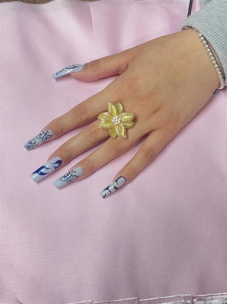 May include: A yellow flower-shaped ring with a clear rhinestone center. The ring is on a finger with light blue nail polish and a blue and white design.