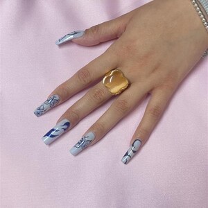 May include: A hand with long, light blue nails with black and white designs. The nails are painted with a flame design. The hand is wearing a gold ring with a large, oval-shaped stone.