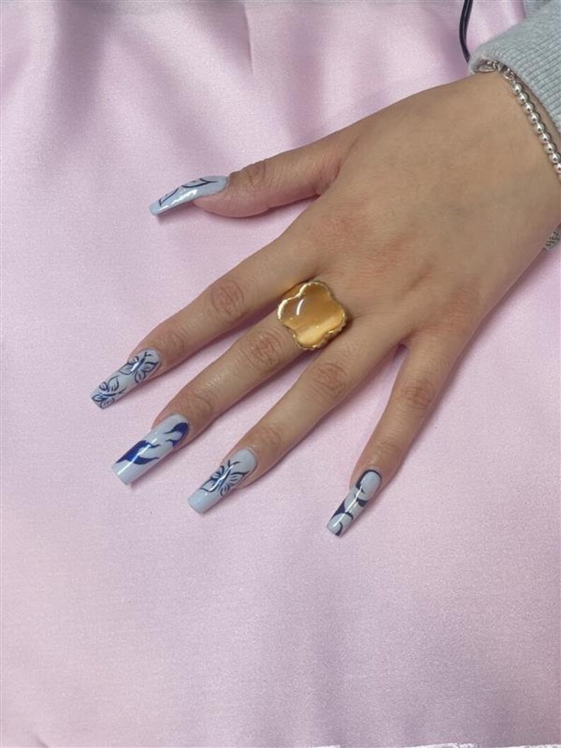 May include: A hand with long, light blue nails with black and white designs. The nails are painted with a flame design. The hand is wearing a gold ring with a large, oval-shaped stone.