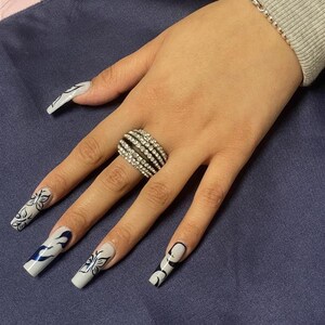 May include: A hand with long, white acrylic nails decorated with black butterfly and flame designs. The hand is wearing a silver ring with multiple rows of clear rhinestones.