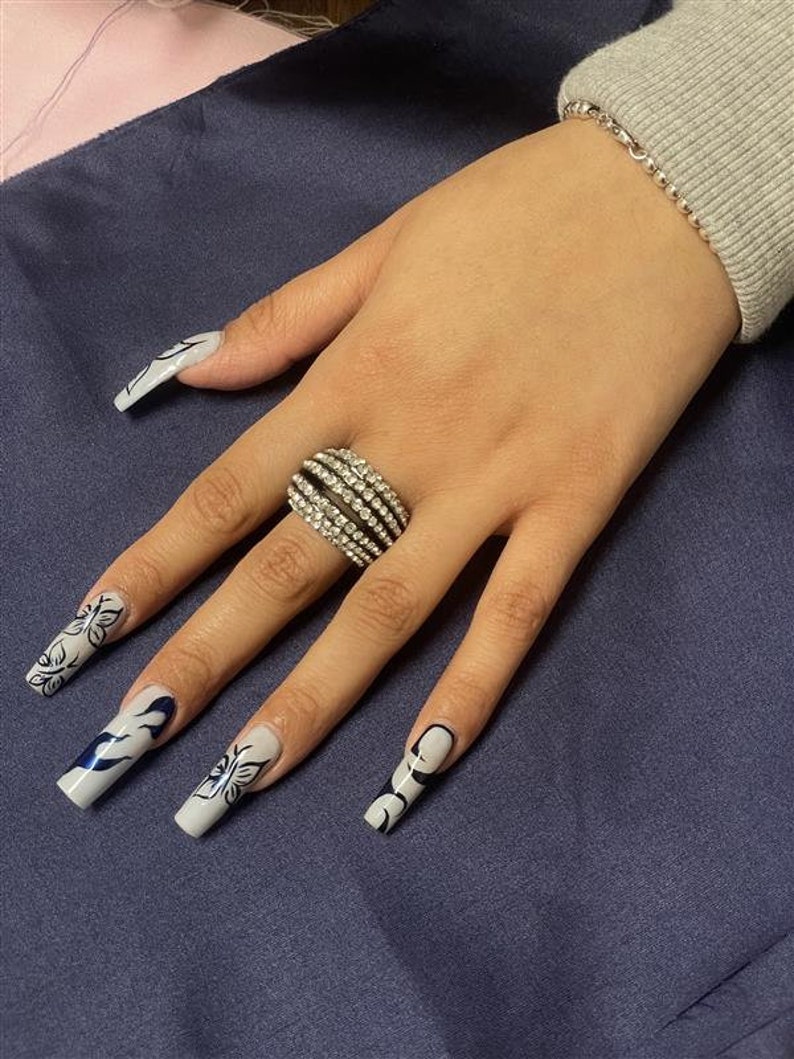 May include: A hand with long, white acrylic nails decorated with black butterfly and flame designs. The hand is wearing a silver ring with multiple rows of clear rhinestones.