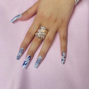 May include: A hand with long, light blue nails with black butterfly and swirl designs. The hand is wearing a gold ring with clear stones.