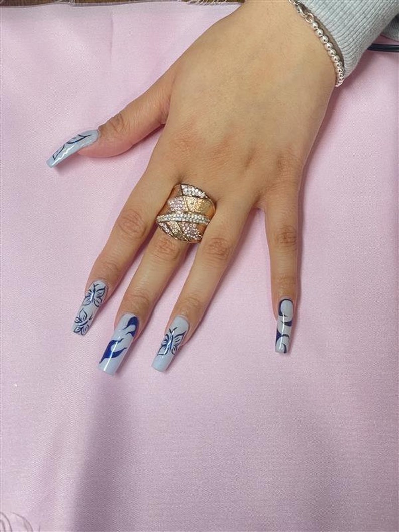 May include: A hand with long, light blue nails with black butterfly and swirl designs. The hand is wearing a gold ring with clear stones.