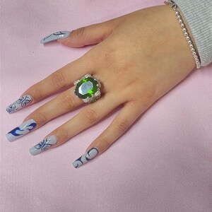 May include: A silver ring with a large, oval-shaped green gemstone surrounded by smaller clear gemstones. The ring is on a hand with long, light blue nails with black and white designs.