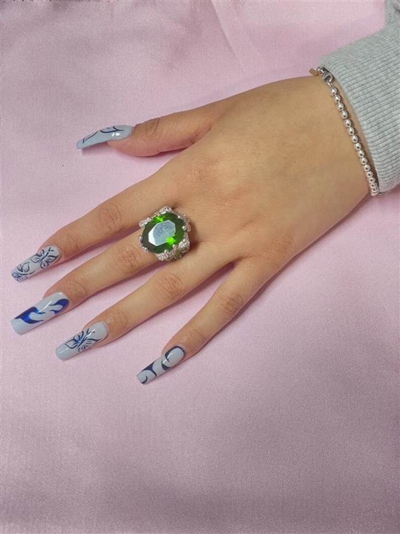 May include: A silver ring with a large, oval-shaped green gemstone surrounded by smaller clear gemstones. The ring is on a hand with long, light blue nails with black and white designs.
