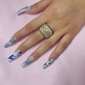 May include: A hand with long, light blue nails with blue butterfly designs. The hand is wearing a gold ring with clear rhinestones.