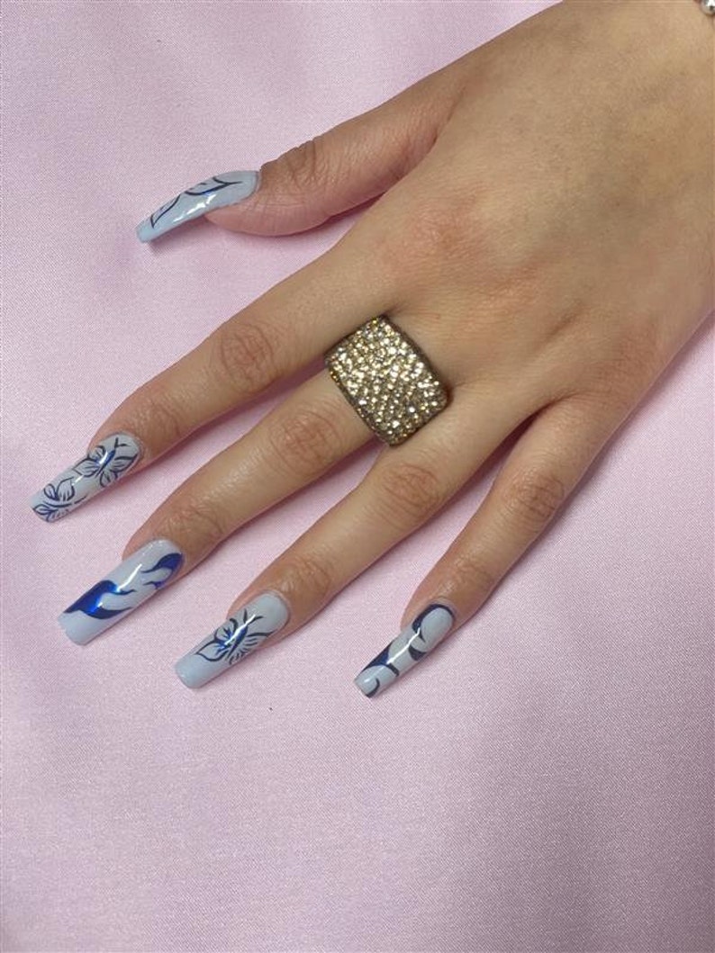 May include: A hand with long, light blue nails with blue butterfly designs. The hand is wearing a gold ring with clear rhinestones.