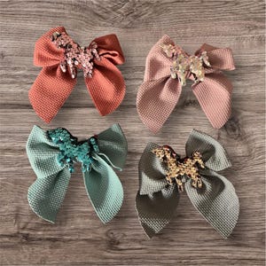 May include: Four fabric hair bows in shades of coral, pink, teal, and gray. Each bow has a textured waffle weave pattern and is embellished with sequins in a horse shape. The bows are arranged on a wooden surface.