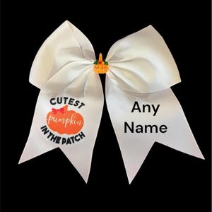 以下が含まれることがあります： 「Cutest pumpkin in the patch」という文字とカボチャのデザインが施された白いヘアボウ。ボウの反対側には「Any Name」という文字が書かれています。