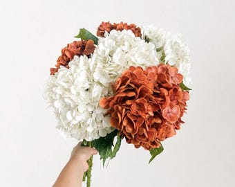 May include: A bouquet of artificial hydrangea flowers featuring large, rounded clusters of petals in alternating shades of crisp white and deep burnt orange, with green stems and leaves visible at the base of the floral arrangement.