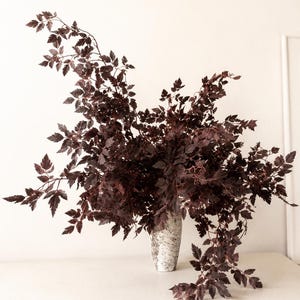 Peut inclure: Un arrangement décoratif de feuilles bordeaux foncé dans un vase blanc moucheté. Les feuilles ont un aspect délicat et superposé, créant un affichage complet et texturé. Le vase est cylindrique et ajoute une touche d'élégance à l'arrangement floral.