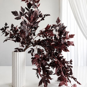 Peut inclure: Un grand vase cylindrique blanc à la surface texturée en chevron contient une grande branche arquée aux feuilles dentelées d'un bordeaux profond. Le feuillage s'étend vers le haut et retombe vers le bas, créant une silhouette asymétrique.