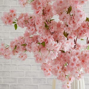 May include: A bouquet of pink artificial cherry blossoms in a white vase. The flowers are arranged in a cascading style, with the branches extending out from the vase.