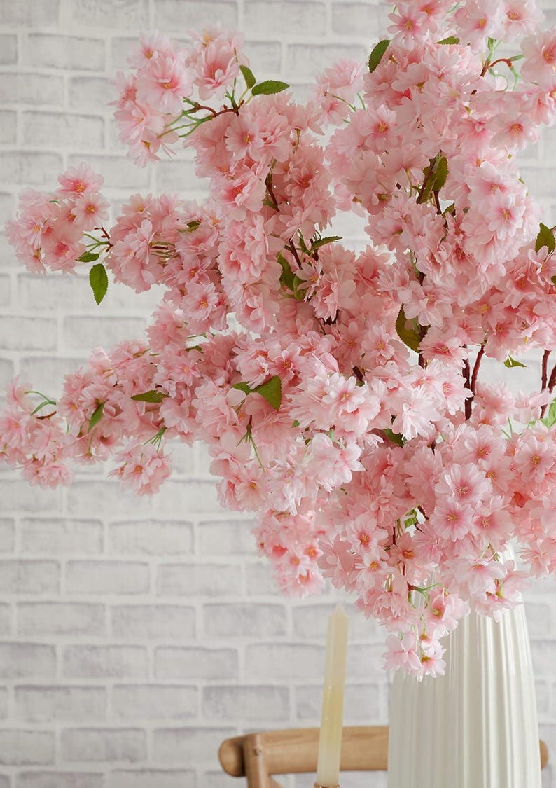 May include: A bouquet of pink artificial cherry blossoms in a white vase. The flowers are arranged in a cascading style, with the branches extending out from the vase.