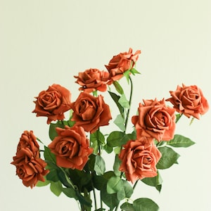 May include: A bouquet of faux roses in shades of burnt orange. The artificial flowers have detailed petals and are surrounded by green leaves. The stems are visible at the bottom, creating a natural arrangement. Perfect for home decor.