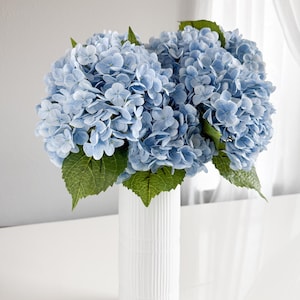 Pode incluir: Um buquê de flores de hortênsia azul claro com folhas verdes em um vaso branco com ranhuras verticais. As flores estão agrupadas, criando uma aparência exuberante. O vaso cilíndrico está sobre uma superfície branca.