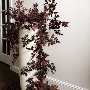 May include: A tall, cylindrical white floor vase with a textured, ribbed surface. The vase is filled with cascading, dark burgundy-coloured foliage featuring small, serrated leaves that drape over the sides and extend outward in multiple directions.