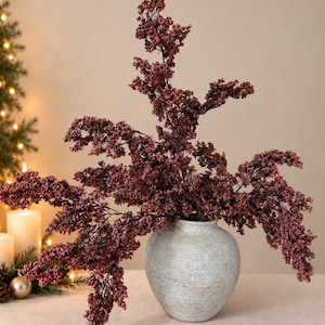 May include: A decorative arrangement of dark red berry-like sprigs displayed in a light grey, horizontally ribbed ceramic vase. The plant stems feature numerous small, textured clusters in a deep burgundy hue, creating a wide, branching shape.