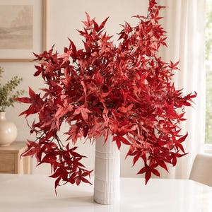 May include: A tall, white, cylindrical vase with a vertical ribbed texture holds a large, dense arrangement of vibrant red maple leaves. The branches spread outwards, creating a full, rounded display of deep crimson foliage with pointed edges.