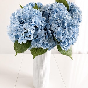 May include: A bouquet of light blue hydrangea flowers with green leaves arranged in a tall, white, cylindrical vase featuring a vertical ribbed texture. The flowers have multiple small, rounded petals and are densely clustered together.