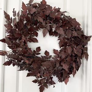 Peut inclure: Une couronne circulaire composée de nombreuses feuilles dentelées brun foncé et de petites grappes de baies assorties. Le feuillage est densément disposé autour d'une ouverture centrale, créant une forme arrondie et pleine d'une couleur brune uniforme.