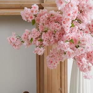May include: A white vase with a pink cherry blossom branch arrangement. The vase is tall and slender with a textured surface. The cherry blossom branch is full of pink flowers and green leaves.