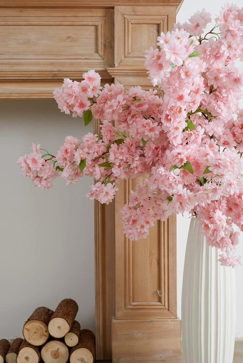May include: A white vase with a pink cherry blossom branch arrangement. The vase is tall and slender with a textured surface. The cherry blossom branch is full of pink flowers and green leaves.