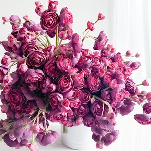Può includere: Un lussureggiante bouquet di fiori di ranuncolo artificiali in varie tonalità di viola e rosa. I fiori sono disposti in un vaso bianco, creando un'elegante esposizione floreale. L'arrangiamento include fiori e boccioli completamente sbocciati.
