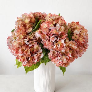 May include: A large bouquet of artificial hydrangea flowers in shades of dusty pink, coral, and muted orange with green leaves. The flowers are arranged in a tall, cylindrical white vase featuring a vertical ribbed or textured surface pattern.