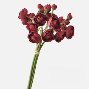 May include: A bouquet of burgundy artificial ranunculus flowers with green stems tied together with a brown ribbon.