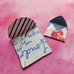 May include: Two small, handmade paper envelopes. One is light green with blue script and a striped flap. The other is white with a floral pattern and a blue polka dot interior. The envelopes are on a pink background.